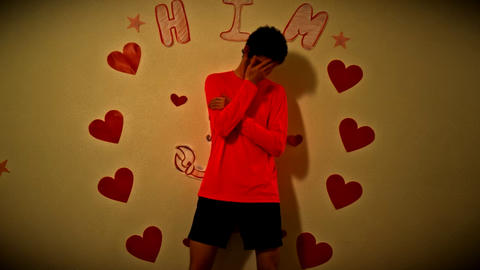 A person in a red long-sleeved shirt and black shorts stands against a beige wall, covering their face with their hand in a gesture suggesting shyness or embarrassment. The wall is decorated with hand-drawn red hearts, yellow stars, and the word 